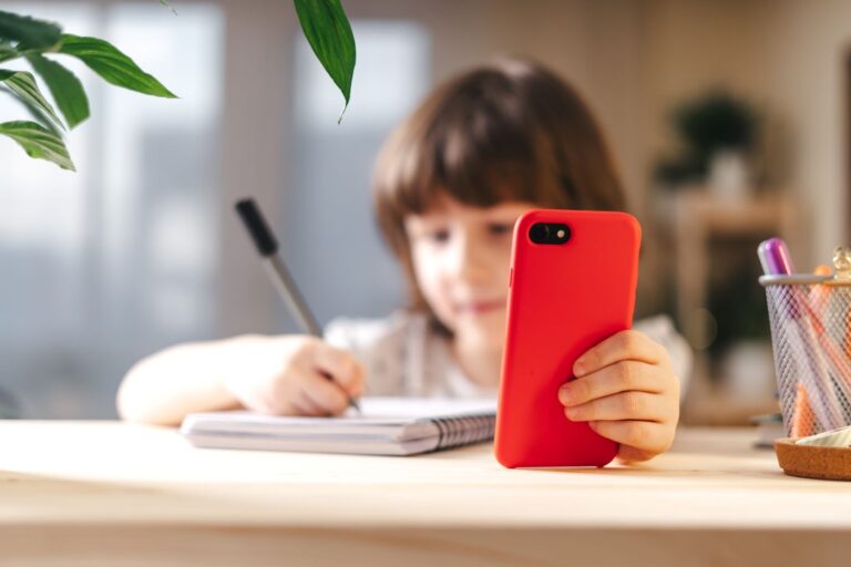 Criança representando a hiperconectividade ao segurar um celular vermelho enquanto escreve em um caderno.